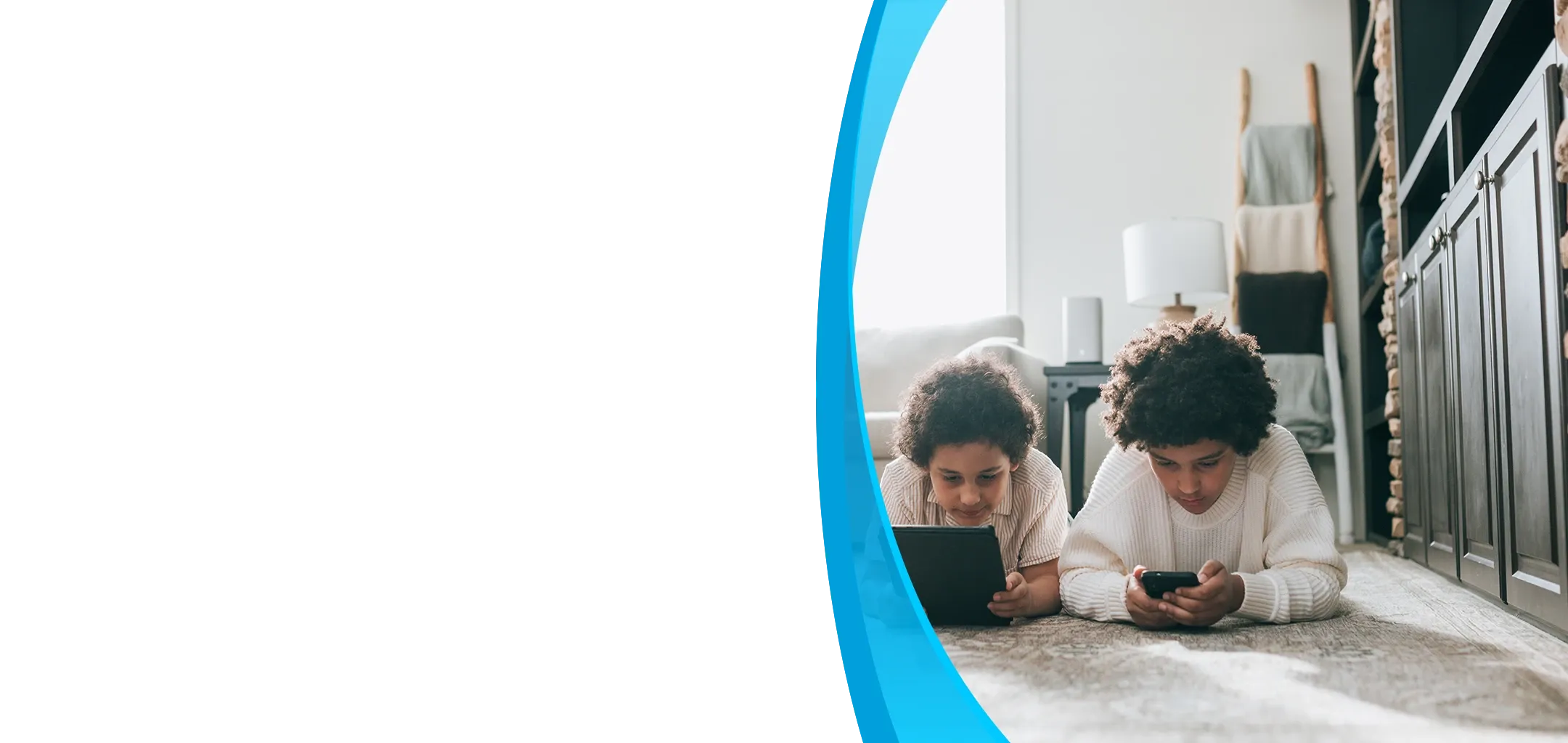 Two curly-haired kids lie on a living room rug using a tablet and phone.